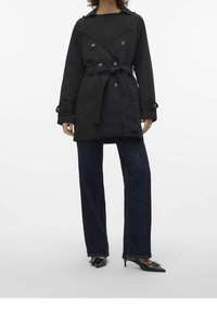 Abrigo negro estilo trench con cierre de botones doble y cinturón en la cintura, combinado con jeans de mezclilla oscura de pierna ancha y zapatos negros puntiagudos.