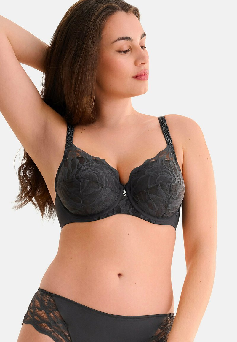 Soutien-gorge en dentelle noire avec motif floral, bonnets rembourrés et bretelles décoratives. Présente un détail de logo argenté au centre et une bande sous lisse.