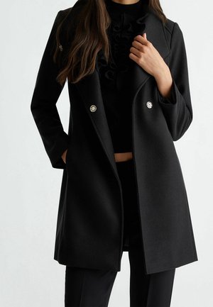 Frau trägt einen schwarzen, doppelt geknöpften Mantel mit goldenen Knöpfen über einem schwarzen, mit Rüschen verzierten Oberteil, eine Hand in der Tasche, die andere hält den Revers des Mantels.