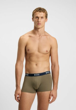 Young man with curly hair wearing olive green boxer briefs with black waistband labeled "BOSS," standing against a plain light background.