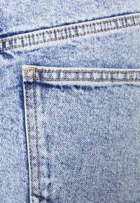 Jeans de denim azul claro com costuras visíveis, dois bolsos e uma textura suave com um ligeiro padrão desbotado.