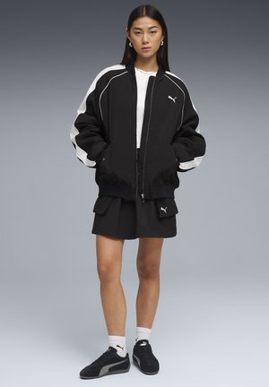 Giovane donna che indossa una giacca Puma bianca e nera, pantaloncini cargo neri, calzini Puma bianchi e scarpe da ginnastica Puma nere, in piedi dritta.