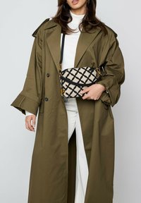 Grüner Trenchcoat mit weiten Ärmeln, kombiniert mit einer gemusterten Umhängetasche mit schwarzem geometrischem Design auf cremefarbenem Hintergrund.