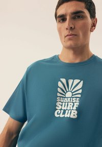 Niebieska bawełniana koszulka z okrągłym dekoltem. Na przodzie znajduje się biały nadruk w postaci słońca oraz tekst "SUNRISE SURF CLUB".