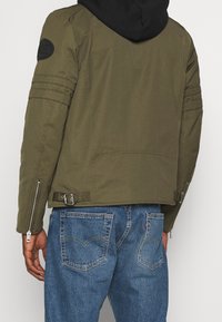 Man som bär en olivgrön jacka med spännband och svart huva, i kombination med blå denimjeans, sedd bakifrån.