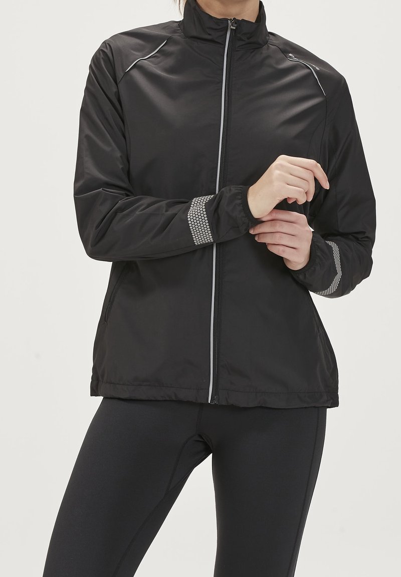 Chaqueta negra con cremallera y cuello alto, detalles reflectantes y rayas texturizadas en las mangas. Material liviano, acabado suave y diseño simple.