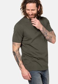 Camiseta de algodón color verde oliva con mangas cortas y cuello redondo. Textura suave, ajuste casual. Tatuajes visibles en el brazo. Combinada con jeans azules.
