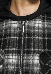 Black and gray plaid zip-up jacket with a soft texture, featuring a silver zipper and a black hood. Notable details include two front pockets.