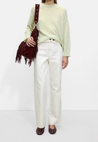 Pull en maille vert clair, pantalon blanc à jambes larges, sac à main bordeaux à franges, et mules marron cloutées avec un bout carré.