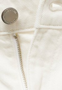 Valkoinen denimpuku näyttää metallisen napin, jossa on brändäys, hopeaketjun ja näkyvät ompelu yksityiskohdat lähellä oleva näkymä.
