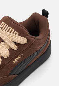 Brown suede sneaker with black accents, beige laces, and gold branding. Features a textured surface and low-profile, cushioned design.