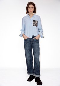 Femme debout, les mains dans les poches, portant une chemise bleu clair avec une poche imprimée léopard et des détails aux poignets, un jean foncé retroussé et des bottes noires.