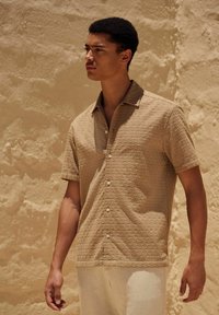 Chemise à manches courtes, à boutons, en tissu texturé beige clair avec un motif subtil, portée avec un pantalon de couleur claire, sur fond de sable.