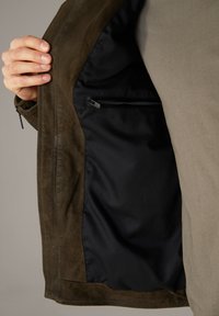 Brun ruskindsjakke med sort indvendigt foer, der viser en lynlåslomme på venstre side og en glat stoftekstur.