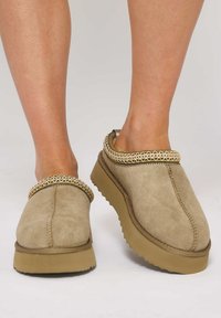 Scarpe slip-on in suede beige con tomaia texturizzata, decorate con una bordatura intrecciata in tonalità oro e una suola in gomma spessa e a contrasto.