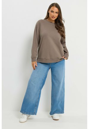 Mujer con cabello largo y castaño, vistiendo una sudadera marrón holgada, pantalones vaqueros de pierna ancha azules y zapatillas blancas, posando frente a un fondo liso.