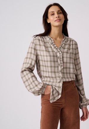 Femme portant un chemisier beige et marron à carreaux avec des volants et des manches longues, associé à un pantalon en velours côtelé marron taille haute, une main dans la poche.