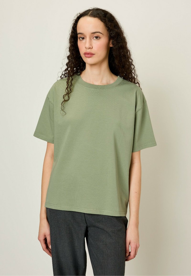 Olīvu zaļš kokvilnas T-krekls ar brīvu piegriezumu, īsām piedurknēm un apaļu kakla izgriezumu; gluda tekstūra ar redzamām šuvju detaļām.