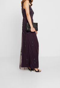 Woman in a sleeveless, floor-length dark purple dress with bead embellishments, holding a black textured clutch and wearing black high-heeled sandals.