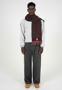 Grå sweatshirt, brun tørklæde med rød tekst og frynser, grå plisserede bukser, brune sko. Modellen står mod en neutral baggrund.