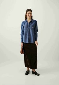 Camisa de ganga com botões e dois bolsos no peito, combinada com uma saia longa castanha e mocassins pretos. O modelo segura uma bolsa castanha.
