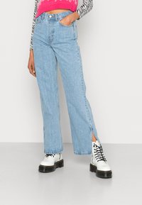 Jeans de cintura alta, de cor azul clara, com pernas largas e fendas laterais, em lavagens desbotadas, combinados com botas de combate brancas e um top cropped rosa.