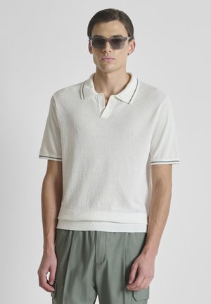 Giovane uomo con occhiali da sole neri, polo bianca a maniche corte e texture, e pantaloni cargo grigi larghi su sfondo neutro.