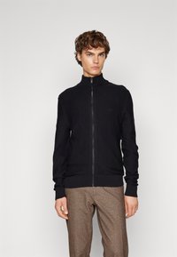 Svart zip-up genser med høy krage, ribbede mansjetter og kant. Discert logo detaljer på brystet og glatt strikket tekstur.