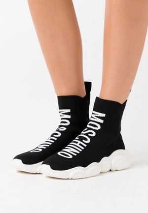 Sorte sokkelignende sneakers med hvid "MOSCHINO" tekst og kraftige hvide såler båret på bare ben mod en hvid baggrund.