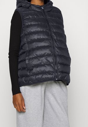 Person iført en sort quiltet ærmeløs hættevest over en sort langærmet top og lysegrå løstsiddende bukser, stående mod en ensfarvet baggrund.