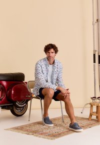 Heren overhemd met bloemenpatroon in lichte stof, gecombineerd met een grijze t-shirt en marineblauwe shorts. Gedragen met grijze schoenen. Vintage rode scooter op de achtergrond.