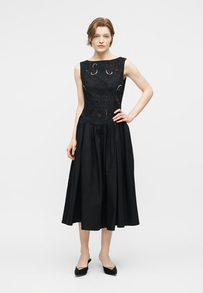 Kobieta stojąca z ręką na biodrze, ubrana w czarną sukienkę bez rękawów z koronkowymi detalami na dopasowanym gorsecie i rozkloszowaną spódnicą midi.