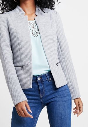 Kvinde iført en lysegrå tekstureret blazer over en lyseblå bluse med blondekant og tætsiddende blå jeans, står med afslappede hænder.