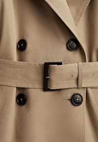 Trench-coat beige à double boutonnage, boutons noirs et ceinture assortie en tissu avec une boucle rectangulaire noire. Texture lisse.