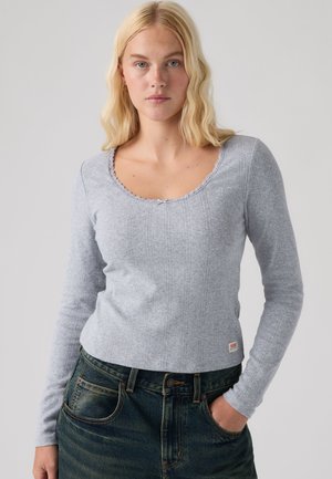 Donna bionda con occhi azzurri indossa un top lavorato a maglia a maniche lunghe grigio chiaro e jeans blu scuro, in piedi con una mano in tasca.