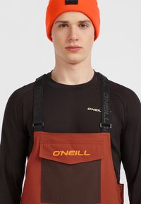 Pantalones de tirantes O'Neill en color naranja quemado y marrón, hechos de tejido sintético; cuentan con tirantes ajustables y un bolsillo frontal con el logo.