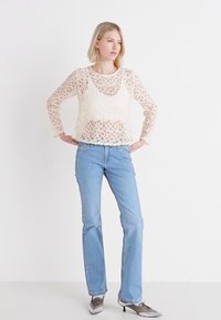 Krajkový top v krémovej farbe s kruhovými vzormi, skombinovaný so svetlomodrými džínmi a striebornými lodičkami s špicatou špičkou. Jednoduché pozadie.
