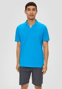 Türkise Poloshirt mit V-Ausschnitt, kurzen Ärmeln und glatter Textur. Kombiniert mit grauen Shorts, vor einem neutralen Hintergrund präsentiert.