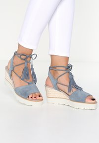 Blue suede wedge sandals featuring a closed heel, open toe, crisscross ankle straps with tassels, and a woven espadrille-style midsole.