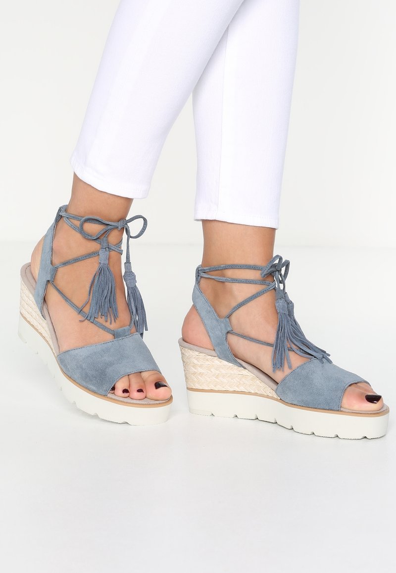 Blue suede wedge sandals featuring a closed heel, open toe, crisscross ankle straps with tassels, and a woven espadrille-style midsole.