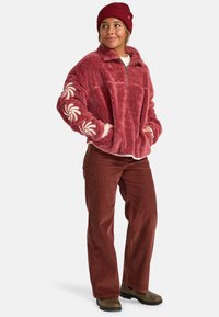 Pullover in pelliccia sintetica rossa con colletto, mezza zip e ricamo floreale bianco sulle maniche. Abbinato a pantaloni in velluto a coste marroni e stivali.