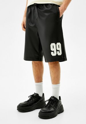 Mand iført sorte sportsnetshorts med hvid "99" på venstre ben, hvide crew strømper og sorte chunky sneakers, hænderne i lommerne.