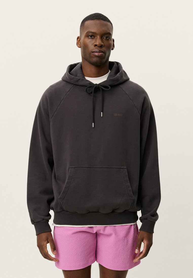 Μαύρο φούτερ με κουκούλα, τσέπη καγκουρό, κορδόνι και ριμπ τελείωμα. Φοράει ροζ κοντό σορτς με υφή από κάτω.