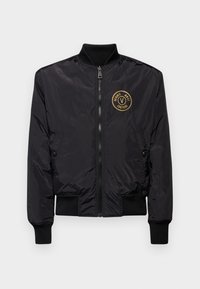 Svart bomberjacka i glänsande nylon, med dragkedja fram, ribbade ärmslut och nederkant, samt en rund guldfärgad logotyp på vänster bröst.