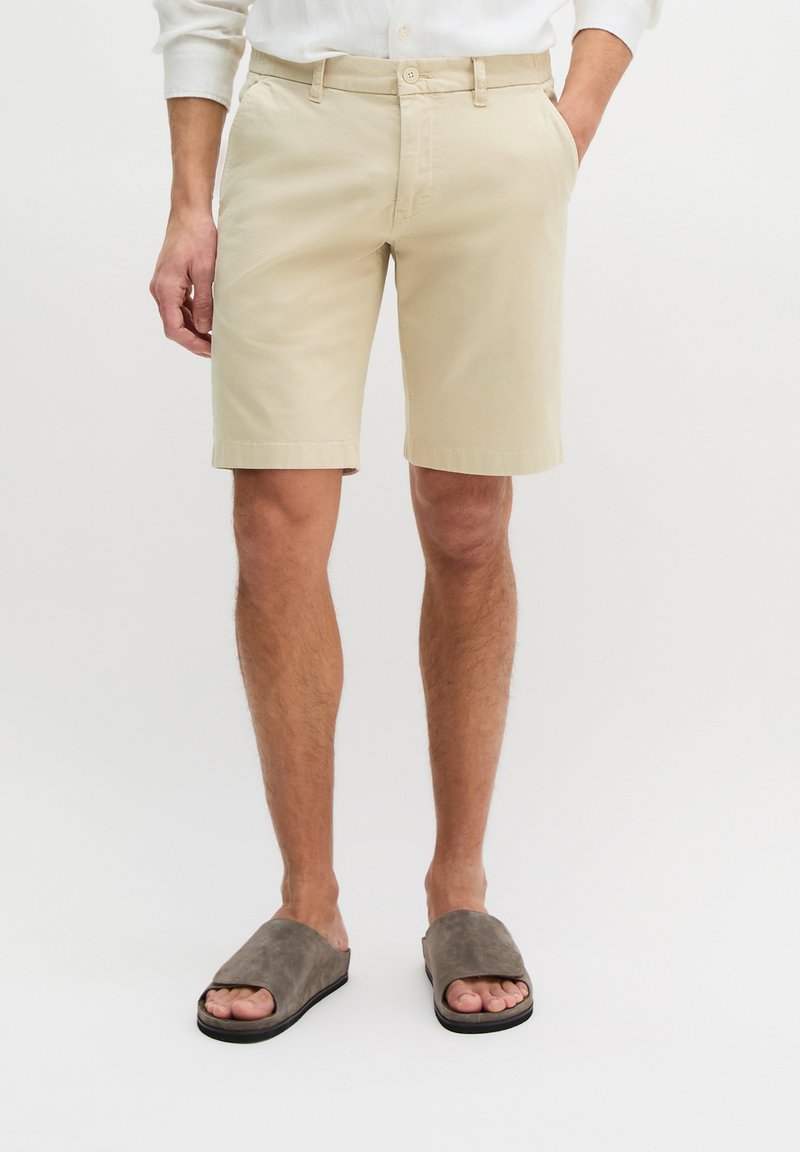 Homme portant un short beige jusqu'aux genoux, une chemise blanche et des sandales grises, debout avec une main dans la poche sur un fond blanc.