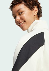 White high-collar jacket with a black panel across the shoulder. Soft texture and modern design, featuring a zip closure at the back.