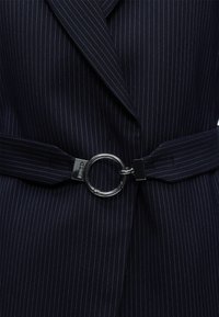 Σκούρο μπλε μπλέιζερ με λεπτή ρίγα, φαρδιές πέτες και ζώνη με μεγάλη ασημένια κυκλική αγκράφα στη μέση.