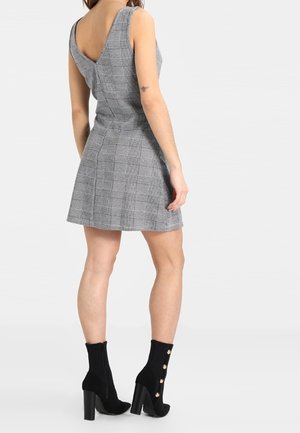Vrouw in een mouwloze grijze geruite jurk met diepe V-rug en zwarte enkellaarsjes met hoge hak en zijknopen, staand met de rug naar de camera.