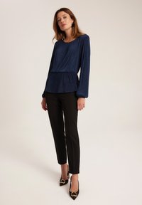 Granatowa bluzka z długim rękawem o teksturowanym wykończeniu i swobodnym kroju, zestawiona z czarnymi dopasowanymi spodniami i wysokimi szpilkami z złotymi akcentami.