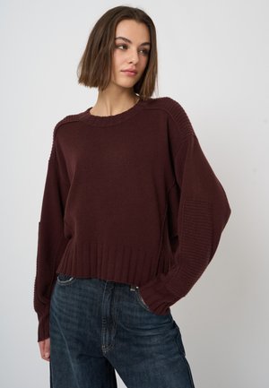 Jonge vrouw met kort bruin haar die een losse donkerbruine gebreide trui en blauwe spijkerbroek draagt, staand tegen een eenvoudige lichte achtergrond.